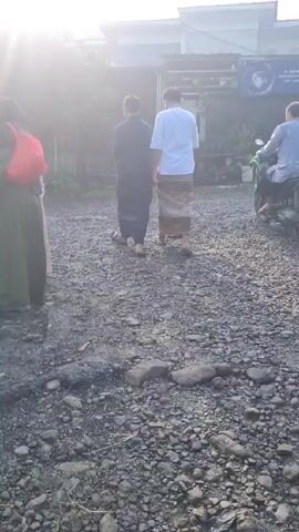 Here is a matter-of-fact description of the video:

The video shows a scene in what appears to be a yard or open space covered with gravel. In the foreground, there are a few larger rocks among the smaller ones. 

At the start, two men are walking away from the camera towards a building in the background. One is wearing a dark, long-sleeved shirt or robe and pants, and the other is in a light-colored, short-sleeved shirt with a patterned sarong. A person on a motorcycle, wearing a long-sleeved shirt, is also seen driving away in the same direction.

Shortly after, a young boy wearing a light blue shirt and dark pants joins the group walking away from the camera. The building in the background has a sign with text and an image, and there are some plants and other items visible near its entrance. The sun is shining brightly, creating glare and obscuring some details of the scene.