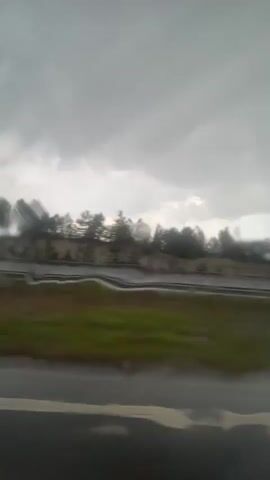 The video depicts a rainy and overcast landscape seen from a moving vehicle. In the foreground, the wet surface of the window is visible, with raindrops distorting the view. The middle ground shows a roadside with green vegetation and a dark asphalt road. Beyond that, there are lines of trees and a blurred background of hills or mountains, obscured by mist or rain. The sky is uniformly gray and cloudy, suggesting a dull, wet day. The motion blur indicates that the vehicle is moving at a moderate speed.