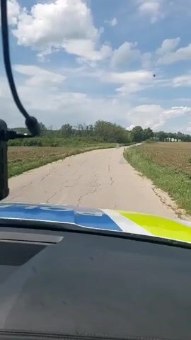 The view from inside a vehicle driving along a rural paved road is captured in the video. The vehicle has a black dashboard with a blue and yellow stripe visible at the top. A device, possibly a microphone or recording device, hangs from the windshield in front of the dashboard. The road stretches ahead, showing cracks and imperfections in the pavement. It is bordered by grassy fields and trees on both sides, under a partially cloudy sky. Police siren noises play in the background.
