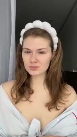 A woman with light skin and long brown hair is shown from the shoulders up in this video. She wears a white headband and a light blue and white striped button-down shirt that is partially unbuttoned. She holds a large black makeup brush with a silver handle. Her face is mostly free of makeup. The background appears to be a room with a white curtain and a window. At 0:03, the image is completely obscured by an opaque white light.