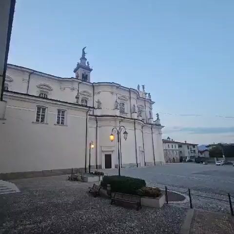 The video shows a view of a building, likely a church, with a tall steeple topped by a statue. The building is light-colored with ornate architectural details and statues of figures along its facade. There are windows and arched openings. In the foreground, there is a paved courtyard area with a few benches and lampposts. The sky is a clear, pale blue, suggesting it might be early morning or late evening. The camera is static for the majority of the clip, with slight movements that provide different angles and close-ups of the building's features, particularly the steeple. There are no visible people or animals in the video.
