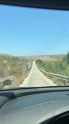 The video shows a view from inside a vehicle looking out through the windshield. A paved road stretches forward into the distance, disappearing over a rise. On either side of the road, there is dry, scrubby vegetation and some rocky terrain. A wire fence runs along the left side of the road. The sky is clear and blue. The overall impression is a rural, possibly arid landscape under a bright sun.