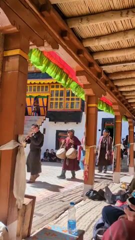 A group of people are walking in procession through an outdoor courtyard. The procession includes individuals wearing traditional clothing, some with hats and others with what appear to be ceremonial headdresses. One person in the front is carrying two drums and appears to be playing them. Another person is dressed in blue and red robes. The procession is moving towards the right side of the frame. In the background, there is a building with yellow-framed windows and colorful banners hanging from an overhang. Some people are seated on the ground on the right side of the frame, observing the procession. The ground is covered with a woven mat or rug. A plastic water bottle is visible on the ground in the foreground. The architecture suggests a location in a mountainous region, possibly a monastery or temple. The lighting indicates it is daytime.