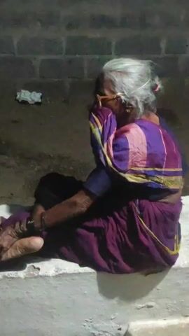 An elderly woman with gray hair and glasses sits on a concrete ledge, wearing a purple sari. She is facing away from the camera, with her right hand covering her face. Her left arm is bent at the elbow, and her legs are crossed. The background appears to be a brick wall.