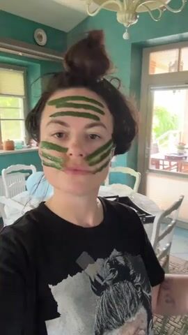 A young woman with dark hair pulled up in a bun, wearing a black t-shirt, is looking at the camera with green stripes of a face mask applied horizontally across her forehead and cheeks. The background appears to be a home interior with turquoise walls, a white chandelier, and furniture. She gestures with her hands near her face as she speaks, and a part of a balloon is visible in the background. The woman is speaking, and the words "you need live content" and "you will get live" can be heard.