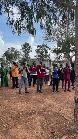 A group of men, some playing brass instruments and drums, are gathered outdoors in a dirt lot. The sky is partly cloudy. Trees and some fencing are visible in the background. The men are wearing various casual outfits and some traditional African attire. The music seems to be in a traditional African style.