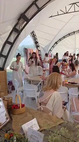 The video shows a gathering of people, mostly women, under a large white tent with exposed metal beams. Many of the attendees are wearing white clothing, including dresses and tops. Some are seated at tables, while others are standing and interacting. In the foreground, a wooden table is visible with a woven basket containing red berries, and some papers or books. Several people in the foreground are partially visible, facing away from the camera, with their backs and shoulders in view. The background shows more people milling around, and some suggest outdoor scenery through open doorways. The overall atmosphere appears to be a casual event or celebration.