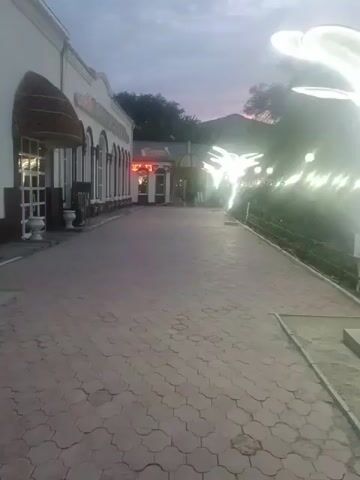 The video shows a paved walkway leading towards a building with arched windows. Large, illuminated flower-like sculptures are prominently displayed along the walkway, casting a bright glow. The sky is dim, suggesting either dawn or dusk. The scene appears to be an outdoor area, possibly a park or the grounds of a restaurant or event venue. There are some trees and greenery visible in the background. The overall atmosphere is serene and visually striking due to the illuminated sculptures. The location is identified as Atyrau, Kazakhstan. No people are clearly visible, and there is no discernible activity taking place other than the static display of the sculptures. The weather appears calm.
