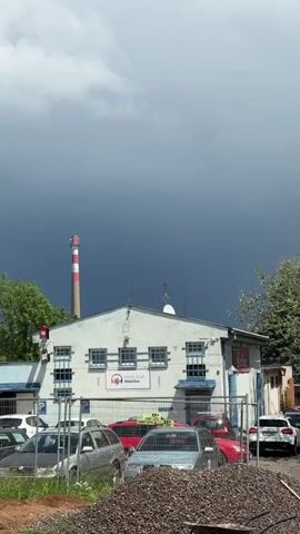 A view of a music club called #bečko with a Coca-Cola sign to the right. Cars are parked in front of the building behind a fence. A smokestack with red and white stripes is visible in the background. The sky is dark with storm clouds.
