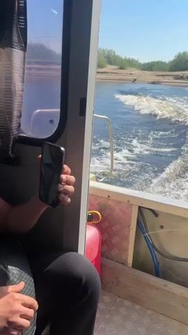 The video shows the rear view from inside a boat traveling on a river. The boat is moving forward, creating a wake in the water. On the left, a person's hand is visible, holding a phone or similar object. The interior of the boat includes a red jerry can and some blue hoses. Outside, the riverbanks are lined with sparse vegetation and sand. The sky is clear and blue. The outboard motor of the boat is visible on the right, with the number 150 on it. The overall impression is of a daytime boat trip on a relatively calm river.