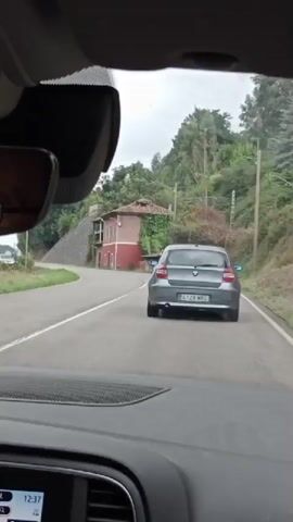 The video, captured from inside a vehicle, shows the rear of a gray hatchback driving on a paved road. The car has a license plate that reads 4128 MRG. To the left of the road, a stone wall runs along the side of a hill covered in vegetation. A reddish-brown building with white trim and dark roof tiles is visible on the right side of the road, partially obscured by greenery. In the distance, trees and foliage cover the hills, and a power line pole is visible. The sky is overcast. The timestamp on the lower left corner of the screen indicates 12:37.