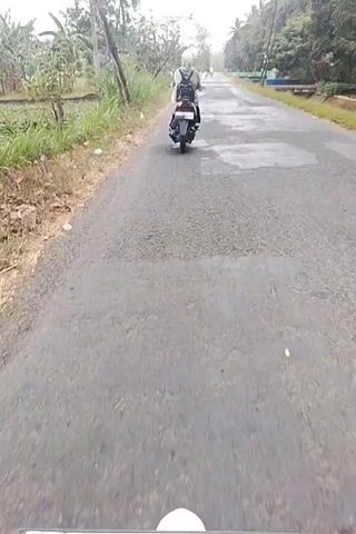 The video shows a person riding a scooter down a paved road. The road is lined with vegetation, including trees and tall grass. The sky is overcast, and the road appears to be wet, possibly from recent rain. The person on the scooter is wearing a helmet and a backpack. The camera is positioned behind the scooter, giving a first-person perspective of the ride.