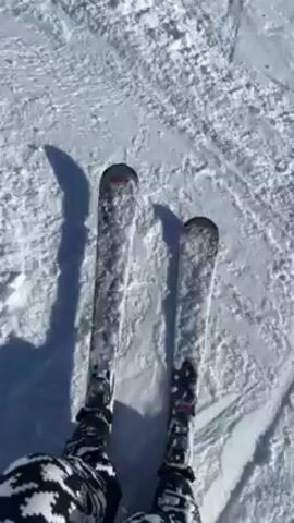 A first-person perspective video shows someone skiing down a snow-covered slope on a bright, sunny day. The camera is fixed on the skier's skis and lower legs, which are clad in black and white patterned ski pants. The skis are white with black and red accents. The snow appears firm and slightly granular, with visible tracks and shadows cast by the skis. The skier is moving at a moderate speed, and the camera angle remains relatively stable, capturing the rhythmic motion of the skis across the snow.