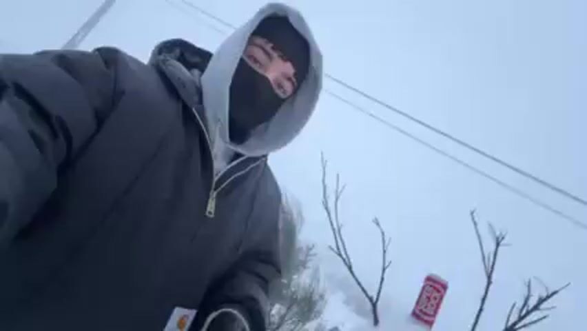 The video shows a person wearing a black balaclava, a grey hooded sweatshirt, and a dark jacket, standing in a snowy environment next to a small snowman. The snowman has twigs for arms, a carrot nose, and a beverage can on its head. The person is holding the camera and appears to be taking a selfie or recording a video. The environment is overcast and snowy, with some visible foliage and power lines in the background.