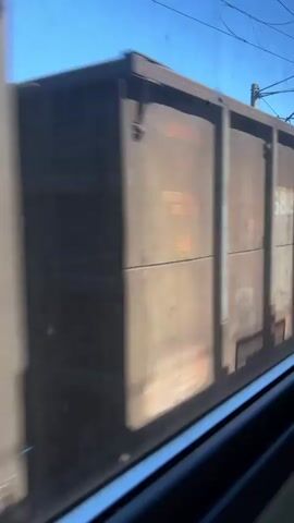 The video shows a train car passing by, seen through a window. The train car is a dark, weathered metal boxcar with vertical seams. There are rectangular panels or markings on the side of the car. Some of these panels are lighter in color, possibly indicating labels or logos. In one section, a logo with a circular design and the word "TRANS" is visible. The train is moving, creating a blur effect in the video. Above the train, power lines and a clear blue sky are visible. The perspective suggests the video was filmed from another moving vehicle, possibly a car or another train, looking out a window.