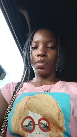 A young Black woman with braided hair and a pink t-shirt with a graphic print of a cartoon face wearing glasses is visible in a selfie video taken from inside a vehicle. She is looking directly at the camera and making various facial expressions, including a pout and a smile. The interior of the car is dark, with a handle visible above her head and a glimpse of the outside through a window.