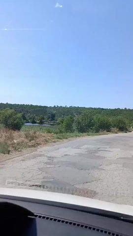 A first-person view from inside a vehicle shows a paved road ahead with patches of disrepair. To the left of the road, there is a body of water with vegetation along its banks. The landscape is characterized by rolling hills covered in trees and bushes under a clear blue sky. The sun is shining, indicating a bright, daytime scene.