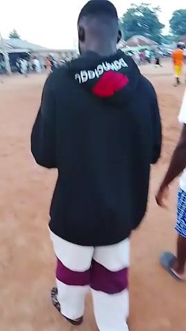 The video shows an outdoor scene with a group of people. The ground appears to be dirt or sand. In the foreground, two people are walking away from the camera. One is wearing a black shirt with a red design and white pants with purple bands. The other is wearing a white t-shirt and blue patterned shorts. In the background, there are more people gathered, some standing and some appearing to be in a group. There are also some buildings and trees visible in the distance. The sky is overcast. The video appears to be shaky and moving.