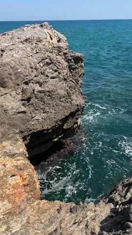 The video shows a rocky coastline with the ocean. The camera is positioned on a rocky outcrop looking towards a larger rock formation in the water. The water is a deep blue-green and appears to be slightly choppy with small waves breaking against the rocks. The sky is clear and blue. There are no people or animals visible in the video. The overall impression is one of a rugged and natural coastal landscape.