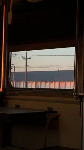 The video shows a view through a train window. Outside the window, a long, low building with many windows or doors is visible, bathed in the warm light of what appears to be sunset or sunrise. Telephone poles and power lines are also visible against a pale sky. The interior of the train car is dark, with a fold-down table in the foreground. The lighting suggests a time of day with low sun angles.