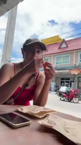 A woman in a red top and baseball cap sits at a table outdoors, eating from a brown paper wrapper. She takes a bite and then adjusts her hair. A phone is on the table in front of her. In the background, there is a street with motorcycles and buildings, including one with a sign that says "Daniel's". The sky is blue with some clouds.