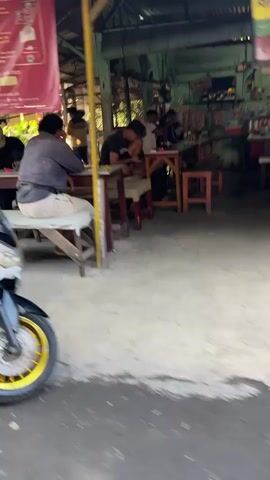 The video shows the exterior of an open-air establishment, possibly a shop or cafe. In the foreground, a portion of a black motorcycle with a yellow rim is visible on the left side of the frame. The ground is a mix of concrete and dirt. In the background, several people are seated at tables. One person in the left-middle ground is wearing a dark jacket and light-colored pants, sitting with their back to the camera. To their right, another person in a dark shirt is leaning over a table. Further back, other individuals are visible, some interacting with each other. The environment appears to be a bustling area, with structures and possibly goods displayed within the establishment. The lighting suggests it is daytime.