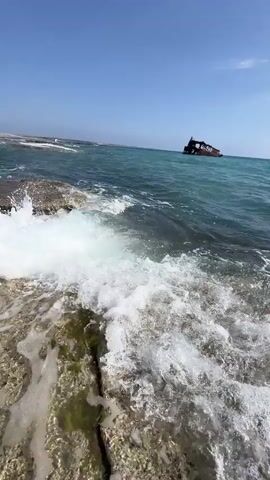 The video shows a close-up view of waves crashing onto rocky shore. In the distance, a shipwreck is visible in the turquoise water under a clear blue sky. The camera is positioned low to the ground, focusing on the action of the waves and the texture of the rocks. The shipwreck appears to be an old, rusty vessel, partially submerged. The overall scene evokes a sense of nature's power and the passage of time.