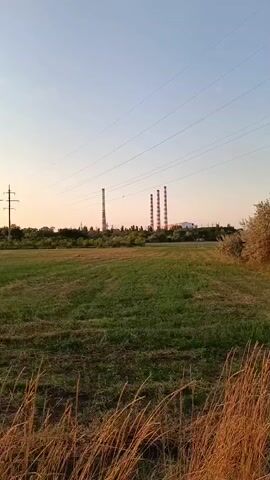 The video shows a wide shot of a rural landscape at sunset. In the distance, there are several industrial chimneys and a power line tower. The foreground is filled with tall, dry grass, and the middle ground is a green field. The sky is a clear, pale blue with hints of orange and pink from the setting sun. The camera pans slowly to the right, revealing more of the power line structure and trees.