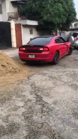 Here is a description of the video.
A red Dodge Charger sits in a driveway facing away from the camera. The car has a black roof and black rims. The license plate is visible and appears to have green and white coloring with black lettering. To the left of the vehicle is a pile of sand. The driveway is made of cracked concrete.  A building, trees, and other cars are visible in the background. The camera slowly zooms in on the back of the red car.