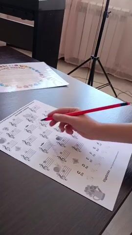 A person's hands are visible as they fill in a worksheet. The worksheet has musical staff lines and a treble clef, with various notes and symbols around it. The person is using a red pencil to write on the paper. In the background, a piano keyboard and a camera tripod are visible. The scene appears to be a music lesson or practice session.