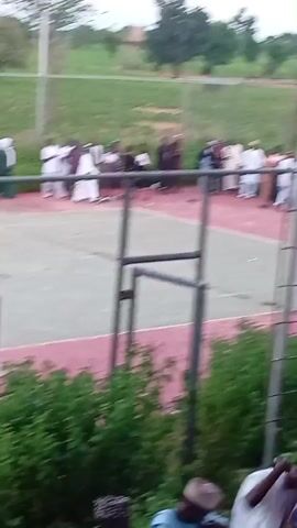 A group of people are gathered on an outdoor court, possibly a tennis court, with a pinkish-red surface and green inner court. There is a chain-link fence surrounding the court, and a tall pole with a light fixture stands within the fenced area. In the foreground, more people are visible, some appearing to be crouching or seated, behind a line of green bushes and a fence. The overall environment appears to be a grassy, outdoor area with trees in the background. The people in the video are dressed in various clothing, with some wearing what appear to be traditional garments. Some individuals are standing, while others are in motion, suggesting some form of gathering or activity is taking place. The lighting suggests it is daytime, possibly late afternoon or early evening. The camera is positioned at a slightly elevated angle, looking down at the scene.
