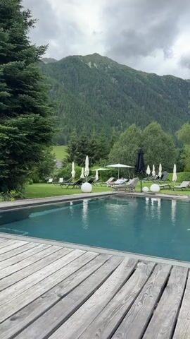 The video begins with a view of an outdoor swimming pool surrounded by a wooden deck. Lush green grass, trees, and a mountain range form the backdrop. Several lounge chairs with umbrellas are placed around the pool. As the video progresses, the camera pans slightly to the right, revealing a building with balconies and windows, and then focuses on a person's legs and feet resting on a lounge chair, indicating someone is relaxing by the pool. The sky is overcast with clouds.