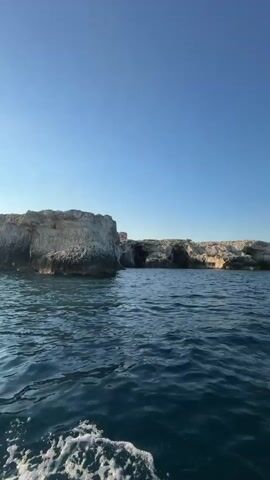 The video captures a boat trip on the Mediterranean Sea, near Syracuse, Italy. The clear blue sky and calm, deep blue water suggest pleasant weather, likely in the morning or late afternoon given the lighting. The boat appears to be a modern passenger vessel, equipped with a white canopy for shade and blue seating areas. Yellow and black hazard tape is visible on the steps leading down into the boat, indicating safety measures.

The initial shot from the water shows a rugged, rocky coastline with caves carved into the cliffs. As the camera pans, it reveals the perspective from the boat, looking towards the open sea and the approaching coastline. A person's bare foot is prominently featured on the deck, suggesting a relaxed and leisurely experience. The boat is moving, creating white foam and ripples in its wake.

There are no people clearly visible engaging in specific activities, but the presence of the boat and the scenic coastal view implies a tour or excursion. The overall mood is serene and picturesque, highlighting the natural beauty of the Italian coastline. Visible text on the boat includes a logo with the text "AQUA MARINE" on both the side of the boat and the steps, indicating the name of the tour operator.