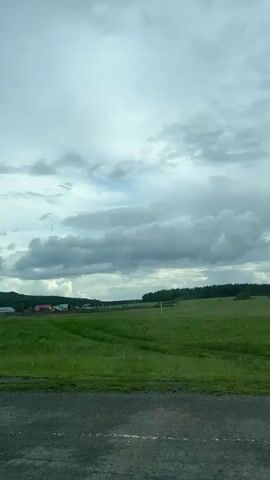 The video shows a rural landscape with a cloudy sky. In the foreground, there is a dirt road or path. Beyond the road, there is a green field with some scattered white flowers. In the middle ground, there is a wooden fence and a small village with several houses, some with red roofs. To the right of the village, there are trees. In the distance, there are more trees and hills. The sky is filled with large, gray clouds. The camera appears to be moving forward, giving a sense of traveling through the scene. There are no people clearly visible in the frame for extended periods.