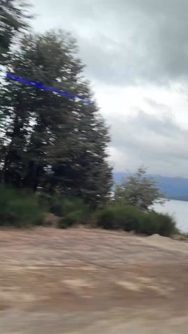 The video shows a view from a moving vehicle of a cloudy sky over a large body of water, possibly a lake, with mountains visible in the distance. The foreground features trees and a dirt road with occasional glimpses of other vehicles and roadside infrastructure.