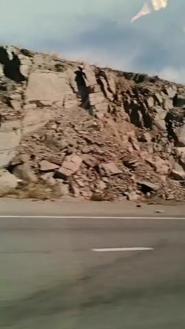 The video shows a side view of a road with a rocky cliff face next to it. The road has a white line painted on it. There is some sparse vegetation at the base of the cliff. The sky is visible above the cliff, with some clouds. The camera appears to be moving.