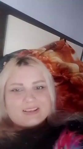 A blonde woman is lying on a bed with a patterned blanket. She is looking at the camera and smiling. The background is out of focus but appears to be a bedroom. The lighting is dim, suggesting it may be nighttime.