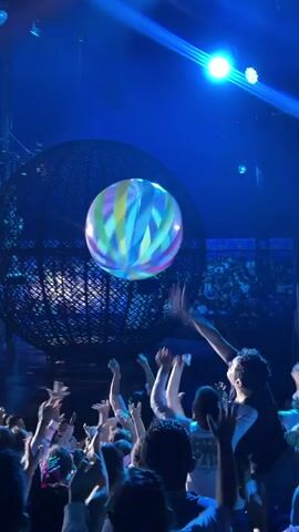 The video captures a concert or live event scene with a large, inflatable beach ball adorned with rainbow stripes being tossed among a crowd. The environment is dimly lit, with bright blue stage lights illuminating the scene, creating a vibrant atmosphere. Several people are visible in the foreground, their hands raised, attempting to interact with the ball. In the background, a large, spherical structure, possibly part of the stage design or a prop, is visible. The overall impression is one of an energetic and engaging audience participation during a performance.