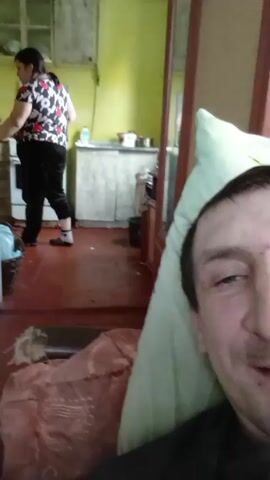 A man lies on a bed, looking at the camera with a smile. Behind him, a woman wearing a patterned shirt and black pants stands in the kitchen, seemingly preparing food. The kitchen is small and somewhat cluttered, with dated cabinets and countertops. A basket sits near the doorway. The overall impression is of a simple, lived-in home.