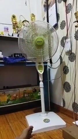 A white standing fan with a green center is in the foreground. The fan is on and its blades are spinning. To the left of the fan, there is a shelf with trophies and a bottle. Below the fan, there is a cabinet with cartoonish images. The floor is made of wood. To the right of the fan, there is a wall with black and white floral wallpaper and several electrical cords. A calendar is hanging on the wall. A person's foot is visible at the bottom left of the frame.
