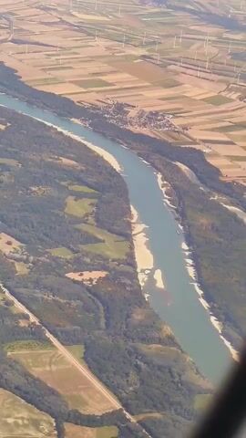 An aerial view shows a wide river flowing through a landscape of fields and trees. The river has sandy banks with some areas appearing shallow or exposed. On the far bank, a small village is visible, nestled among the fields. In the distance, numerous wind turbines are spread across the agricultural land, suggesting a rural or semi-rural setting. The scene appears to be captured during daylight, with the sun casting shadows and highlighting the textures of the terrain. The perspective suggests the viewpoint is from an aircraft.
