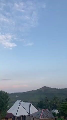 This vertical video captures a serene outdoor scene in Igbara, Nigeria, likely during the late afternoon or early evening. The sky is a soft, pale blue, with wispy white clouds scattered across the upper portion. A prominent hill, covered in lush green vegetation, dominates the mid-ground, with a few scattered buildings visible on its slopes. In the foreground, the white tarp of a tent is visible, suggesting a temporary shelter or event setup. The overall atmosphere is peaceful and tranquil. The sunlight appears gentle, indicating a time of day when the sun is not at its highest point. The presence of a hill and abundant greenery suggests a rural or semi-rural setting. There is no visible text in the video.