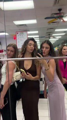 Three young women pose for a selfie in a mirror. The woman on the left wears a white top, black skirt, and white purse. The woman in the middle wears a brown dress. The woman on the right wears a light purple dress. They all have long hair and are holding smartphones. The background shows a diner-like environment with tile floors, a menu board, and other people.