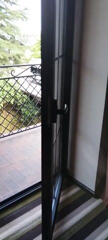 The video pans across a view from what appears to be a window or balcony. The foreground shows a metal railing with a diamond pattern, made of black bars. Below the railing is a brick structure, and to the right of that, a tiled surface. Through the railing, a body of water, likely the sea, is visible under a cloudy sky. Green trees and vegetation are also present, some with palm leaves. Electrical wires are visible in the sky. The overall impression is an outdoor scene with a view of the sea, viewed from a protected area.