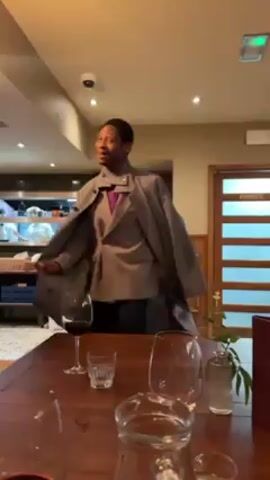 A person dressed in a gray coat and suit enters a restaurant and approaches a partially set table. The table has wine glasses, water glasses, and a small vase with a sprig of greenery. A glass of red wine is on the table. The person puts their coat down on the table and then enters a doorway marked "PRIVATE" behind the table. A server or employee enters the doorway after the first person. The restaurant has a bar area visible with espresso machines, shelves of ingredients, and dish-washing area.