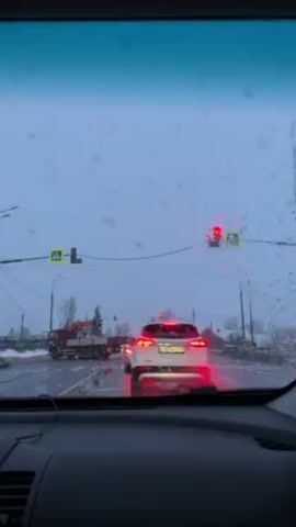 The video shows a view through the windshield of a car stopped at a traffic light on a snowy, overcast day. A white car is directly in front, and a red crane truck is visible further ahead.  Other vehicles are also present on the road. The traffic light is red. Snow is falling.  The windshield is splattered with droplets. Overhead power lines are visible.