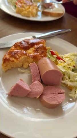 The video shows two plates of food on a wooden table. The plate in the foreground holds a slice of baked casserole, sliced sausage, and a small amount of shredded cabbage salad with red and yellow bell pepper. A knife rests on the plate. The plate in the background has a similar meal and a slice of bread. A person uses a fork and knife to eat from the plate in the background. A small bowl of dark sauce is visible to the left of the plate in the background.