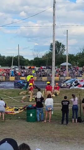 A demo derby event is underway as multiple individuals in bright yellow shirts and helmets, likely event staff or safety personnel, attend to a damaged car. Some are positioned around the vehicle, while others appear to be communicating. Spectators are visible in the background, seated in bleachers or standing, watching the event. The ground is dirt or grass, with some debris and possibly water or mud present in the derby area. A white fence separates the spectators from the derby activity. The sky is partly cloudy.