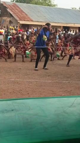 A person in a blue shirt dances in front of a group of children in red-patterned skirts, holding small green and white flags. Behind them, a crowd watches under a tin-roofed building. The person in the blue shirt dances energetically, seemingly leading the children. To the side, a man in shorts watches. In the foreground, a green table with a bottle of water sits, out of focus. The scene appears to be a cultural performance in an outdoor, potentially rural, setting.