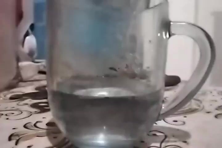A transparent glass mug filled with clear liquid sits on a patterned surface. A stream of clear liquid is being poured into the mug from a jug or pitcher that is not fully visible. The liquid in the mug is rising, indicating the process of filling.